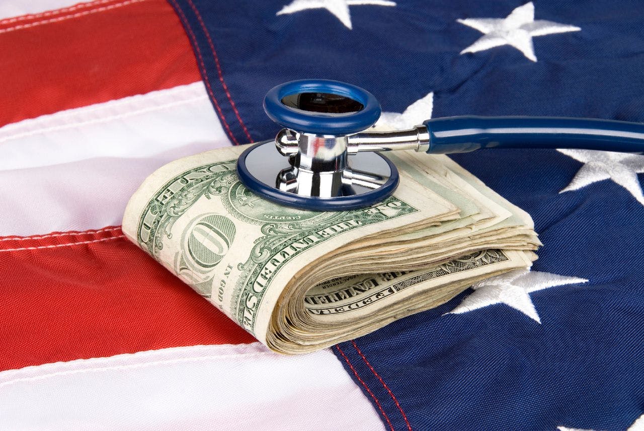 stethoscope on a pack of rolled bills, with an American flag backdrop.