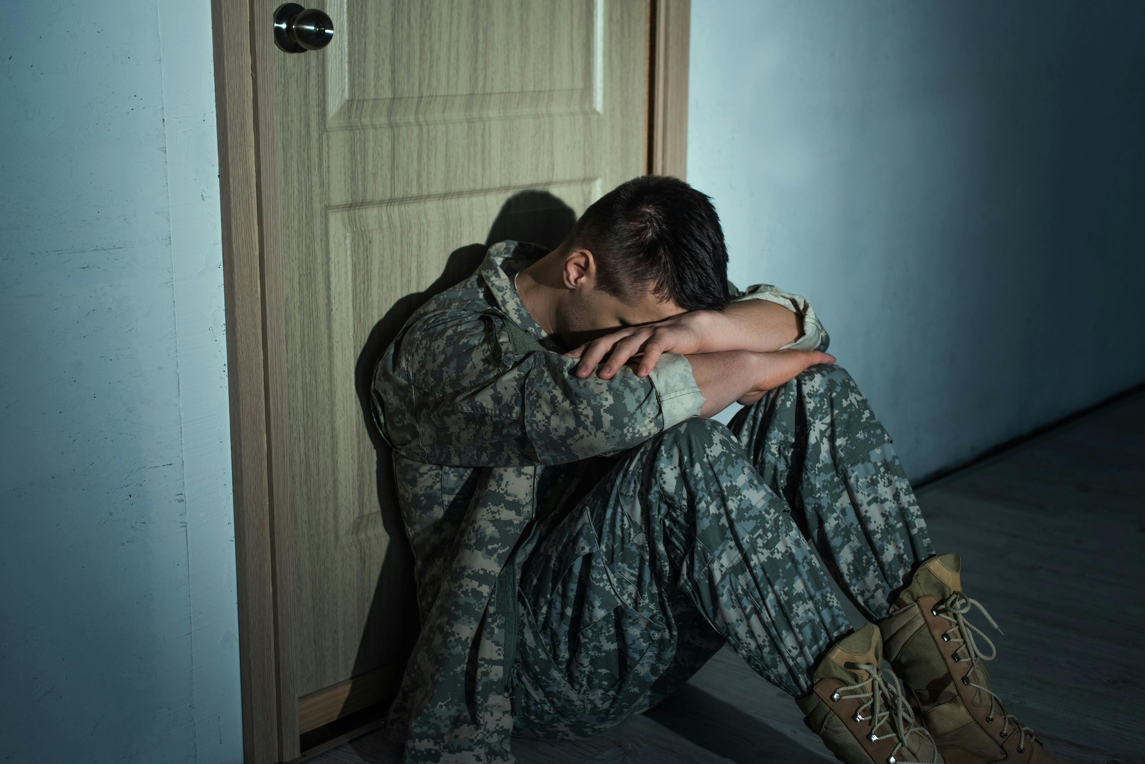 Military man with emotional distress sitting near door in hallway at home at night | Image Credit: LIGHTFIELD STUDIOS - stock.adobe.com